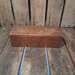 May include: A rectangular wooden storage box with a dark brown finish and visible wood grain. The box features a hinged lid construction with visible finger joints on the side and a small, centered metal latch mechanism on the front panel.