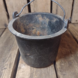 May include: A small, black cast iron pot with a curved metal handle. The pot has a rounded body and a wide opening. The pot is sitting on a wooden surface. The pot is likely used for cooking or display.