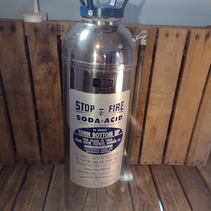 May include: A vintage soda-acid fire extinguisher. The cylindrical metal container has a blue handle and a label with instructions. The label reads "STOP-FIRE SODA-ACID FIRE EXTINGUISHER". It is designed to extinguish fires of wood, paper, textiles, rubbish, and electrical fires.