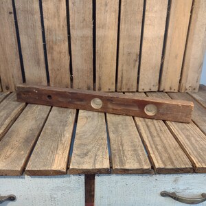 May include: A vintage wooden level with two circular viewing windows. The level is made of dark brown wood and shows signs of wear. It rests on a wooden surface with a rustic, weathered appearance, suggesting it's an antique tool.