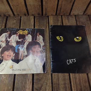 May include: Two vintage vinyl record albums. One album cover features a group of people in white suits with the text "The Osmonds IN CONCERT". The other album cover is black with yellow cat eyes and the word "CATS".