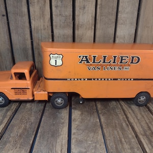 May include: An orange metal toy semi-truck with a trailer featuring the Allied Van Lines logo, the text Allied Van Lines, Inc., and the slogan World's Largest Mover. The truck has black wheels and a shield-shaped emblem with the number 1.