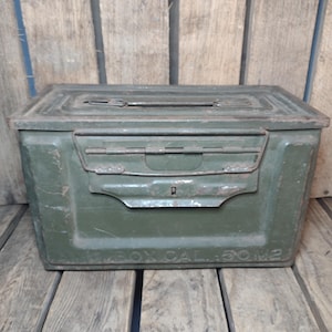 May include: An olive green metal ammunition box with a hinged lid and a latch closure. The box has a rectangular shape with rounded corners and is sitting on a wooden surface. The text "BOX CAL. 50 M2" is visible on the front.