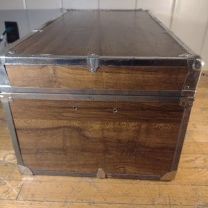 May include: A vintage wooden storage trunk with metal accents. The trunk has a brown wood finish and silver metal trim. The trunk has two metal clasps on the top and two metal hinges on the side.