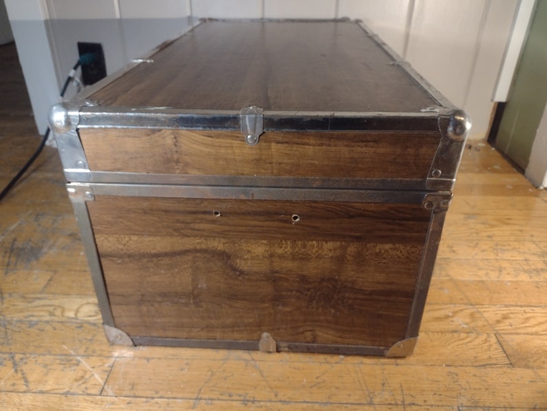 May include: A vintage wooden storage trunk with metal accents. The trunk has a brown wood finish and silver metal trim. The trunk has two metal clasps on the top and two metal hinges on the side.