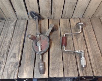 Puede incluir: Un taladro manual vintage con un engranaje circular grande y un mango de madera, junto a una herramienta de berbiquí de metal con una empuñadura giratoria de madera. Ambas herramientas manuales están hechas de acero y madera con piezas mecánicas.