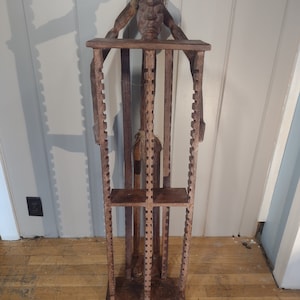 May include: A wooden sculpture of a person with a carved face, standing inside a wooden frame with multiple shelves. The sculpture is made of dark brown wood and has a rustic appearance.