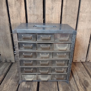May include: A vintage metal parts cabinet with a dark gray finish. It features a rectangular shape with multiple small drawers, each with a clear plastic front and a metal pull. The cabinet is set against a wooden background.