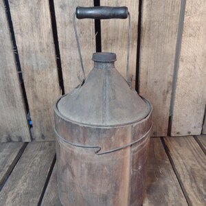 May include: A vintage cylindrical wooden container with a metal top and a central spout. The vessel features a wire handle with a dark wooden grip, secured by metal brackets. The exterior shows a weathered, aged texture with visible wood grain.