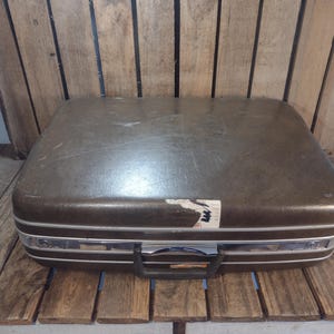 May include: A vintage brown suitcase with silver trim and a black handle. The suitcase has a rectangular shape and a slightly worn appearance, with a partially torn sticker on the top. The suitcase is placed on a wooden surface.