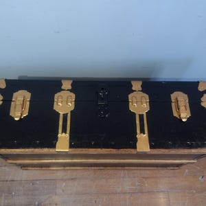 Puede incluir: Un ba&uacute;l de almacenamiento de madera negro con detalles y cierres de metal dorado. El ba&uacute;l tiene una forma rectangular y est&aacute; cerrado.