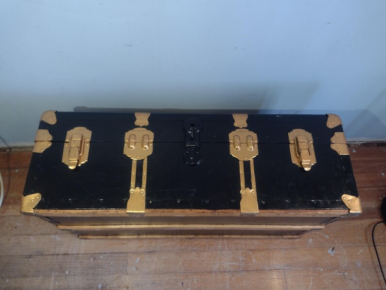 Puede incluir: Un ba&uacute;l de almacenamiento de madera negro con detalles y cierres de metal dorado. El ba&uacute;l tiene una forma rectangular y est&aacute; cerrado.