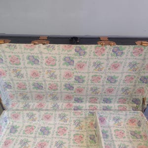 Puede incluir: Una maleta vintage negra con herrajes de lat&oacute;n y un forro con estampado floral. La maleta tiene una tapa con bisagras y un compartimento separado.
