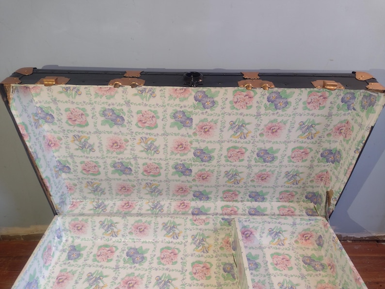 Puede incluir: Una maleta vintage negra con herrajes de lat&oacute;n y un forro con estampado floral. La maleta tiene una tapa con bisagras y un compartimento separado.