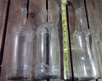 May include: Three clear glass bottles with different textures and designs. The bottles are arranged in a row on a wooden surface. A yellow measuring tape is positioned between the middle two bottles.