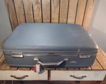 May include: A vintage blue suitcase with silver hardware and a handle. The suitcase has a rectangular shape and is sitting on a wooden surface. A small tag with red, white, and blue stripes hangs from the handle.