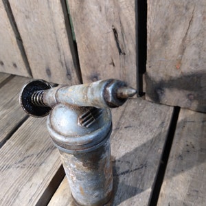 May include: A vintage, silver-colored grease gun with a black handle and a nozzle. The gun is sitting on a wooden surface.