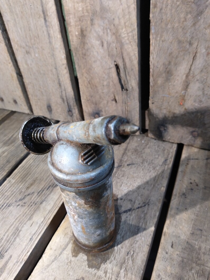 May include: A vintage, silver-colored grease gun with a black handle and a nozzle. The gun is sitting on a wooden surface.