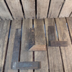 May include: An assortment of vintage metal and wood carpenter's squares and a metal level are arranged on a weathered wooden surface. The tools display signs of age and use, with a rustic aesthetic.