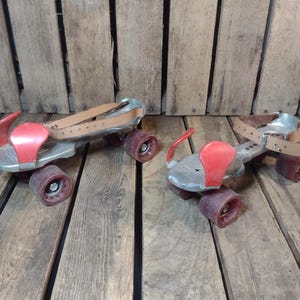 May include: A pair of vintage roller skates with silver metal frames, red toe stops, and brown leather straps. The wheels are a dark reddish-brown color. The skates are displayed on a weathered wooden surface.
