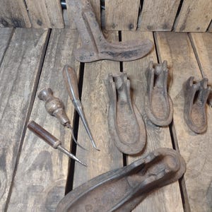 May include: Collection of antique cobbler tools, including shoe lasts and various hand tools. The tools are made of metal and wood, with a rustic, aged appearance. The arrangement includes a shoe form, awls, and other tools.