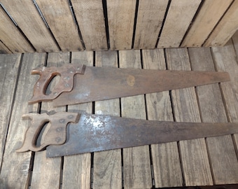 May include: Two antique hand saws with wooden handles and rusty blades are displayed on a wooden surface. The saws are positioned horizontally, showcasing their aged appearance and the intricate teeth along the blades. The handles are a warm brown color.