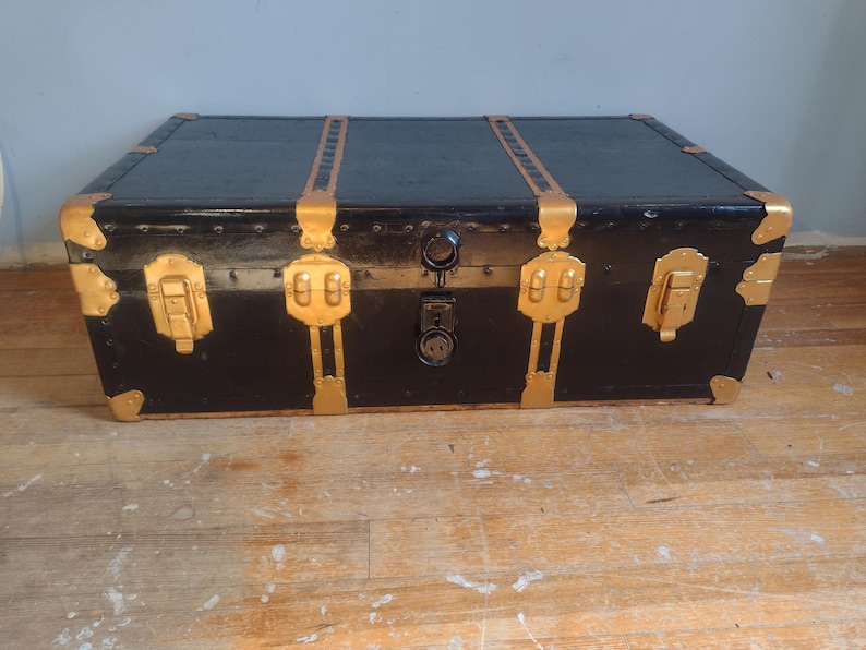 Puede incluir: Un ba&uacute;l vintage negro con detalles dorados y herrajes de lat&oacute;n. El ba&uacute;l tiene dos pestillos a cada lado y una cerradura en el centro.