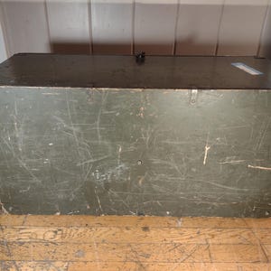 May include: A vintage green wooden storage chest with metal clasps and a worn surface. The chest has a dark green paint finish and shows signs of age and use.