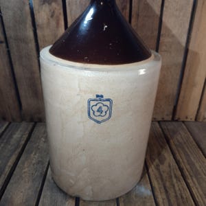 May include: A vintage stoneware jug with a dark brown, cone-shaped top and a beige body. A blue emblem with the number 4 is on the front. The jug is likely used for storing liquids and has a rustic, antique appearance.