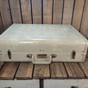 May include: Cream-colored vintage suitcase with gold-toned hardware, including latches and a handle. The suitcase is rectangular with rounded corners and sits on a wooden surface. The suitcase has a combination lock.