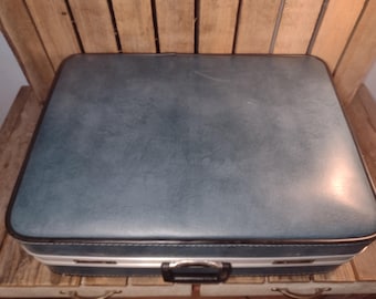May include: Vintage blue suitcase with a black handle and trim. The suitcase has a rectangular shape and is sitting on a wooden surface. The suitcase is a classic travel accessory.