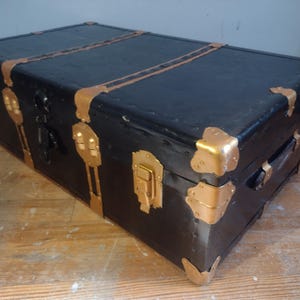 Puede incluir: Un ba&uacute;l vintage negro con detalles y herrajes de metal dorado. El ba&uacute;l tiene una tapa con bisagras y una cerradura.