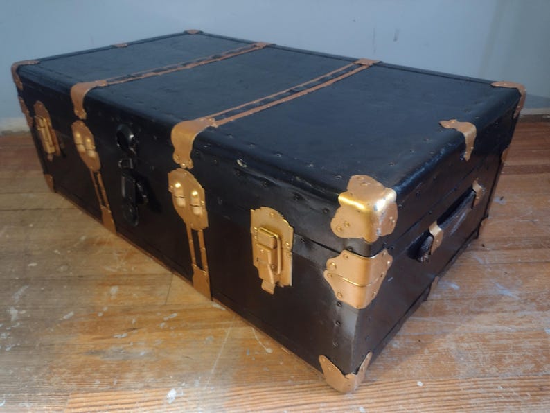 Puede incluir: Un ba&uacute;l vintage negro con detalles y herrajes de metal dorado. El ba&uacute;l tiene una tapa con bisagras y una cerradura.