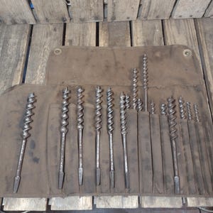 May include: A set of vintage auger drill bits in varying sizes, displayed in a brown canvas tool roll. The bits are made of metal and have a spiral design. The tool roll is open, revealing the bits.