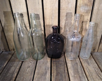 May include: Six glass bottles of various shapes and sizes, including a dark brown bottle, on a wooden surface.