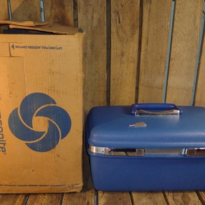 May include: A blue Samsonite hard-sided travel case with a silver latch and handle. The case is sitting on a wooden surface and has a cardboard box behind it. The box is brown with blue text and a blue logo. The text on the box reads "Samsonite" and "Lift and pull across carton".