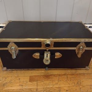 May include: A black trunk with gold-colored metal trim and hardware. The trunk has a rectangular shape with a flat top and is secured with a lock and latches. The trunk is sitting on a wooden floor.