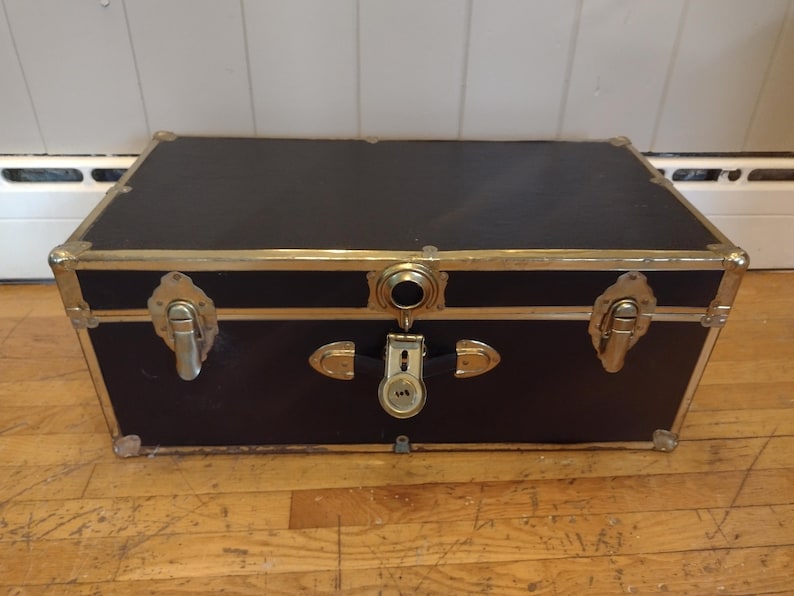 May include: A black trunk with gold-colored metal trim and hardware. The trunk has a rectangular shape with a flat top and is secured with a lock and latches. The trunk is sitting on a wooden floor.