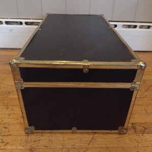 May include: A black trunk with gold-colored metal trim and corner protectors. The trunk has a rectangular shape and is sitting on a wooden floor. The trunk has a hinged lid and a clasp closure. The trunk is likely used for storage.