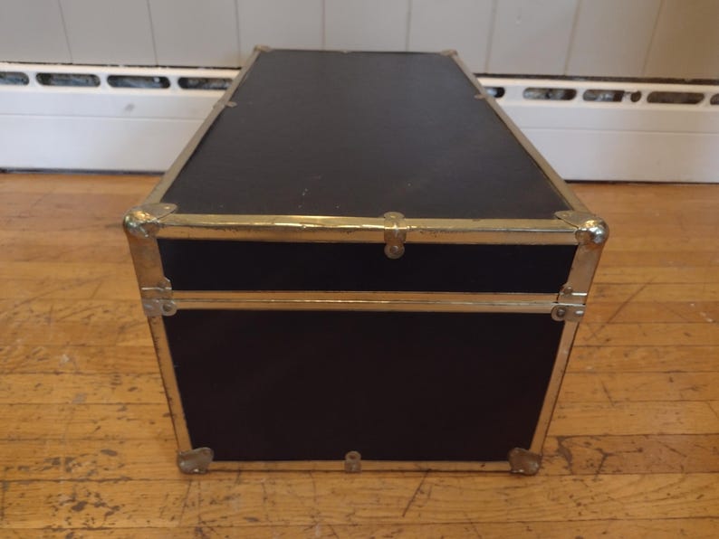 May include: A black trunk with gold-colored metal trim and corner protectors. The trunk has a rectangular shape and is sitting on a wooden floor. The trunk has a hinged lid and a clasp closure. The trunk is likely used for storage.