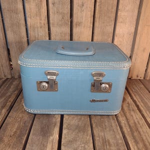 May include: A vintage, sky-blue suitcase with a top handle and two metal latches. The suitcase has a rectangular shape with rounded corners and white stitching. The metal latches and handle are silver-toned. The suitcase is sitting on a wooden surface.