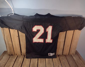 May include: Black football jersey with the number 21 in white with red trim. The jersey is displayed on a wooden surface. The jersey has a Champion logo on the bottom.