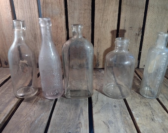 May include: Five clear glass bottles of various shapes and sizes, arranged in a row on a wooden surface.