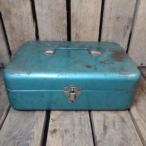 May include: A vintage teal metal toolbox with a handle and a latch. The rectangular box shows signs of wear, with rust spots adding to its aged appearance. The toolbox is set against a backdrop of weathered wooden planks.
