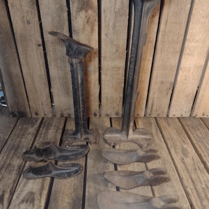 May include: An assortment of antique shoe-making tools, including two shoe lasts and several shoe forms. The tools are made of dark metal and sit on a wooden surface. The background is a wooden wall.