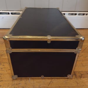 May include: A black trunk with brass-colored metal trim and corner protectors. The trunk has a rectangular shape and is sitting on a wooden floor. The trunk's lid is closed and has a latch.