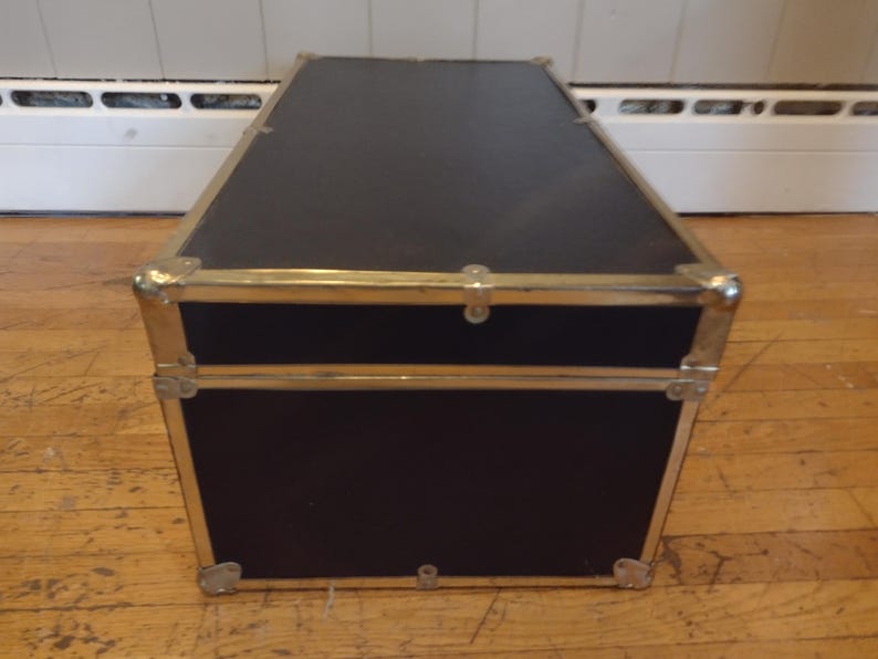May include: A black trunk with brass-colored metal trim and corner protectors. The trunk has a rectangular shape and is sitting on a wooden floor. The trunk's lid is closed and has a latch.