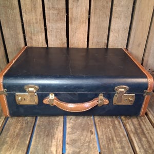 May include: A rectangular suitcase featuring a dark navy blue exterior with tan leather-colored trim along the edges. The suitcase includes a central tan leather-colored handle and two brass-toned metal latches with keyholes on the front panel.