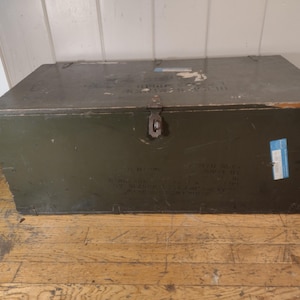 May include: A vintage green wooden storage chest with a metal latch and a blue label. The chest has a worn, distressed look and is sitting on a wooden floor.