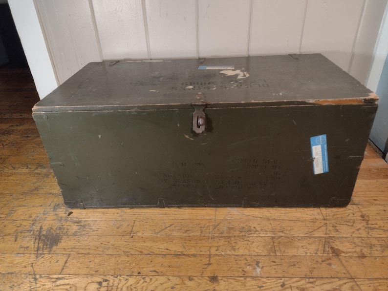 May include: A vintage green wooden storage chest with a metal latch and a blue label. The chest has a worn, distressed look and is sitting on a wooden floor.